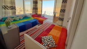 A bedroom with a large bed, a smaller bed, and a desk with a plant. There are curtains and a view of the city. - Jordan Guest House (Wadi Musa)