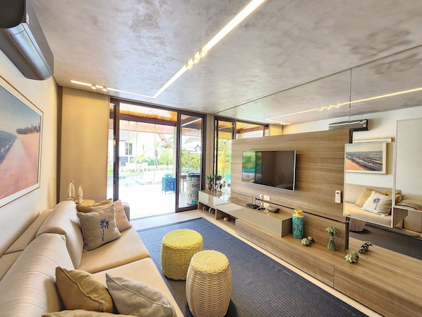 A modern living room with a large flat-screen TV, comfortable seating, and a view of the outdoors. - Bangalo com Piscina Privativa no Malawi Beach Porto de Galinhas (Ipojuca)