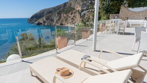 Un espace de détente extérieur baigné de lumière, avec des meubles en osier blancs, une balustrade en verre et une vue sur la mer.