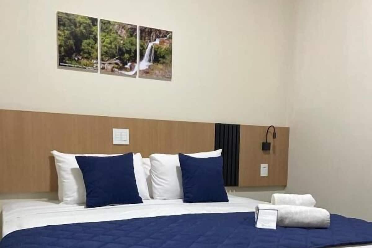 A hotel room with a large bed, white and blue bedding, a wooden headboard, and two framed pictures on the wall.