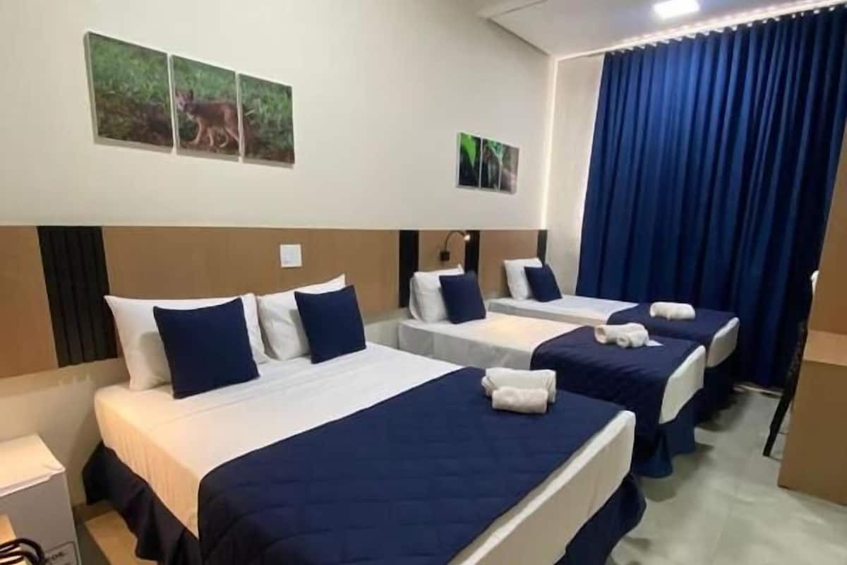 A hotel room with three beds, each with a blue bedspread and white pillows. There are two wall-mounted picture frames, a ceiling light, and a window with blue curtains.
