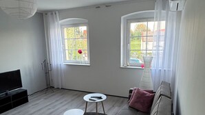 Un salon moderne avec un canapé gris, une petite table ronde et un meuble pour la télévision. Il y a deux fenêtres avec des rideaux, un luminaire blanc au plafond et un radiateur.