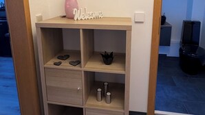 Une petite pièce, de taille modeste, avec un bureau en bois, un vase rose et une pancarte « Bienvenue ». Elle contient trois tiroirs et deux étagères sur lesquelles sont posés des objets cylindriques.