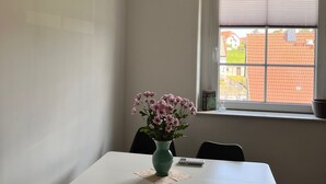 Une pièce avec une table blanche, des chaises noires, un vase vert avec des fleurs, une fenêtre avec des stores et un appareil de climatisation fixé au mur.