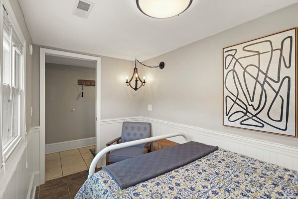 Une chambre à coucher avec une couvre-lit à motifs, une chaise, un lustre et un grand tableau abstrait accroché au mur.