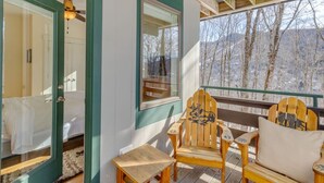 Eine Holzterrasse mit zwei Adirondack-Stühlen und einem kleinen Tisch, eine Glastür, die zu einem Raum führt, und eine Aussicht auf Bäume und Berge.