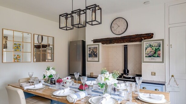 Un comedor con una larga mesa de madera dispuesta para seis personas, un reloj en la pared y obras de arte enmarcadas.