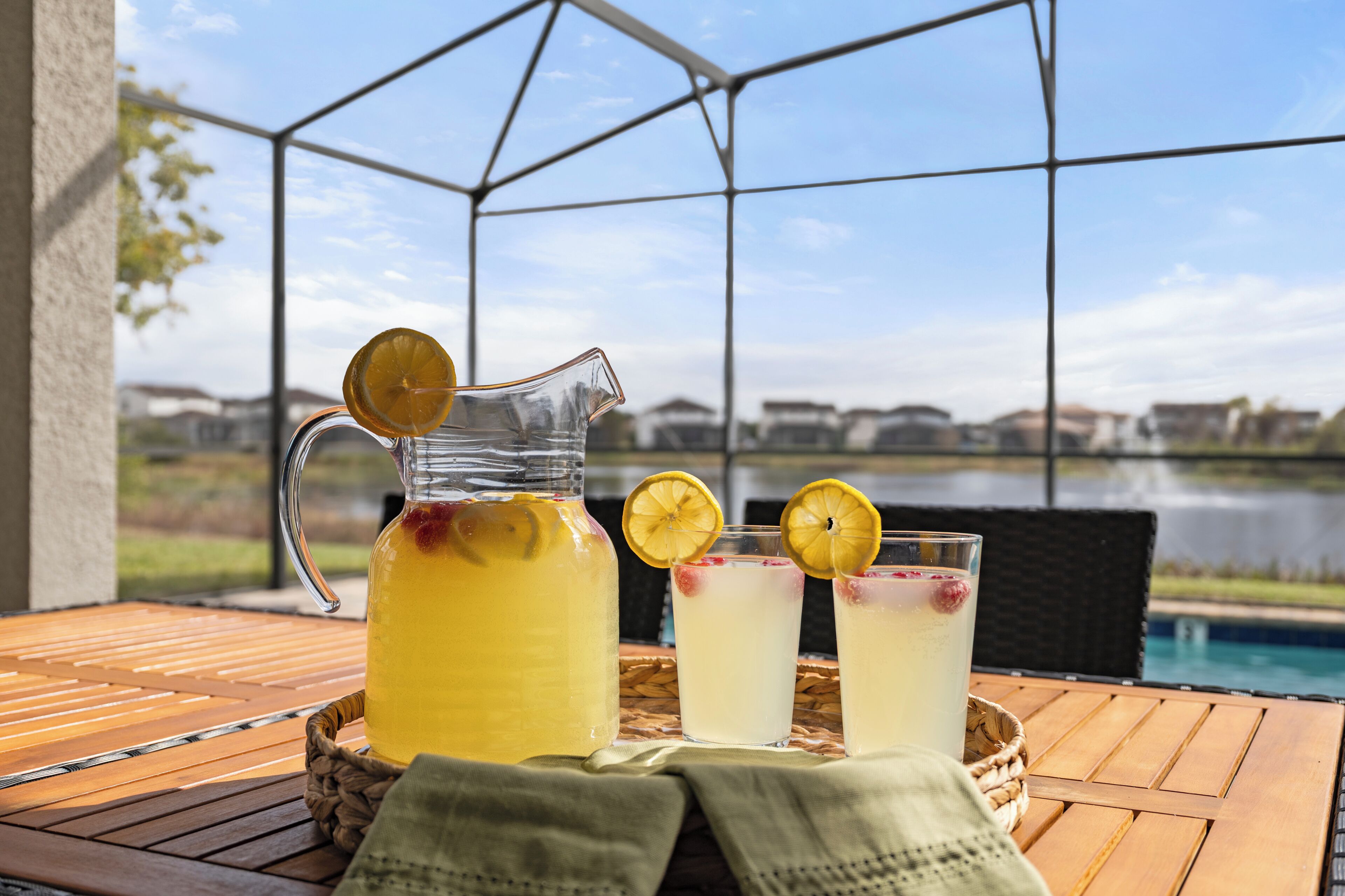 Een houten tafel met een karaf en glazen limonade, op een balkon met uitzicht op een meer en huizen.