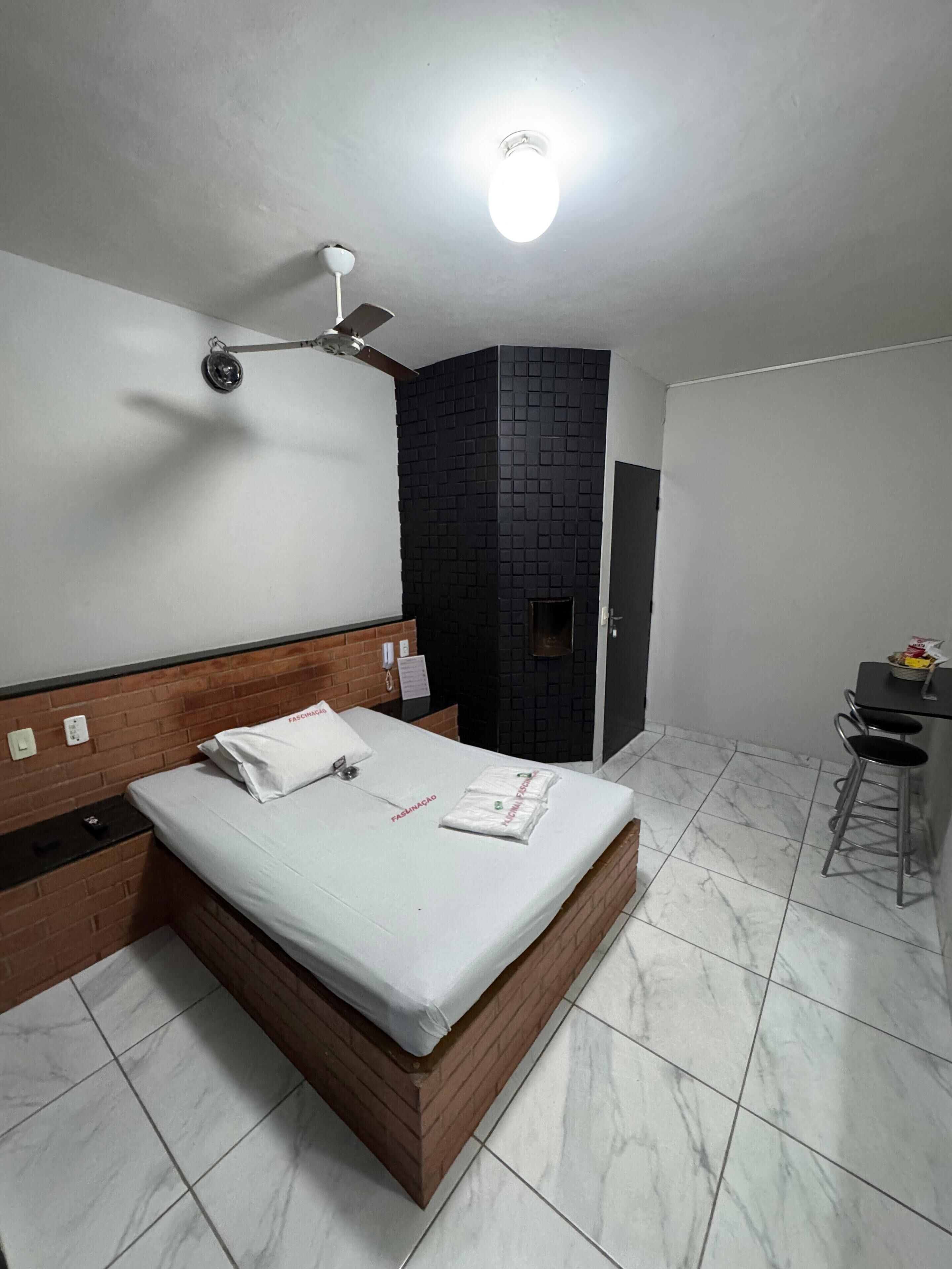 A modern hotel room with a wooden headboard, a bed with white linens, a black accent wall, a ceiling fan, and a small desk with a chair.