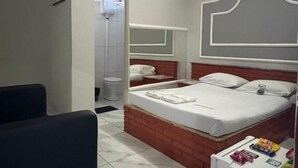 A compact hotel room with a single bed, a small desk, a chair, and a refrigerator. - Motel Fascinação (Contagem)