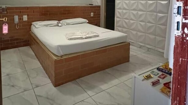 A bedroom with a bed, a brick wall, a white textured wall, a ceiling with a geometric design, and a white refrigerator. - Motel Fascinação (Contagem)