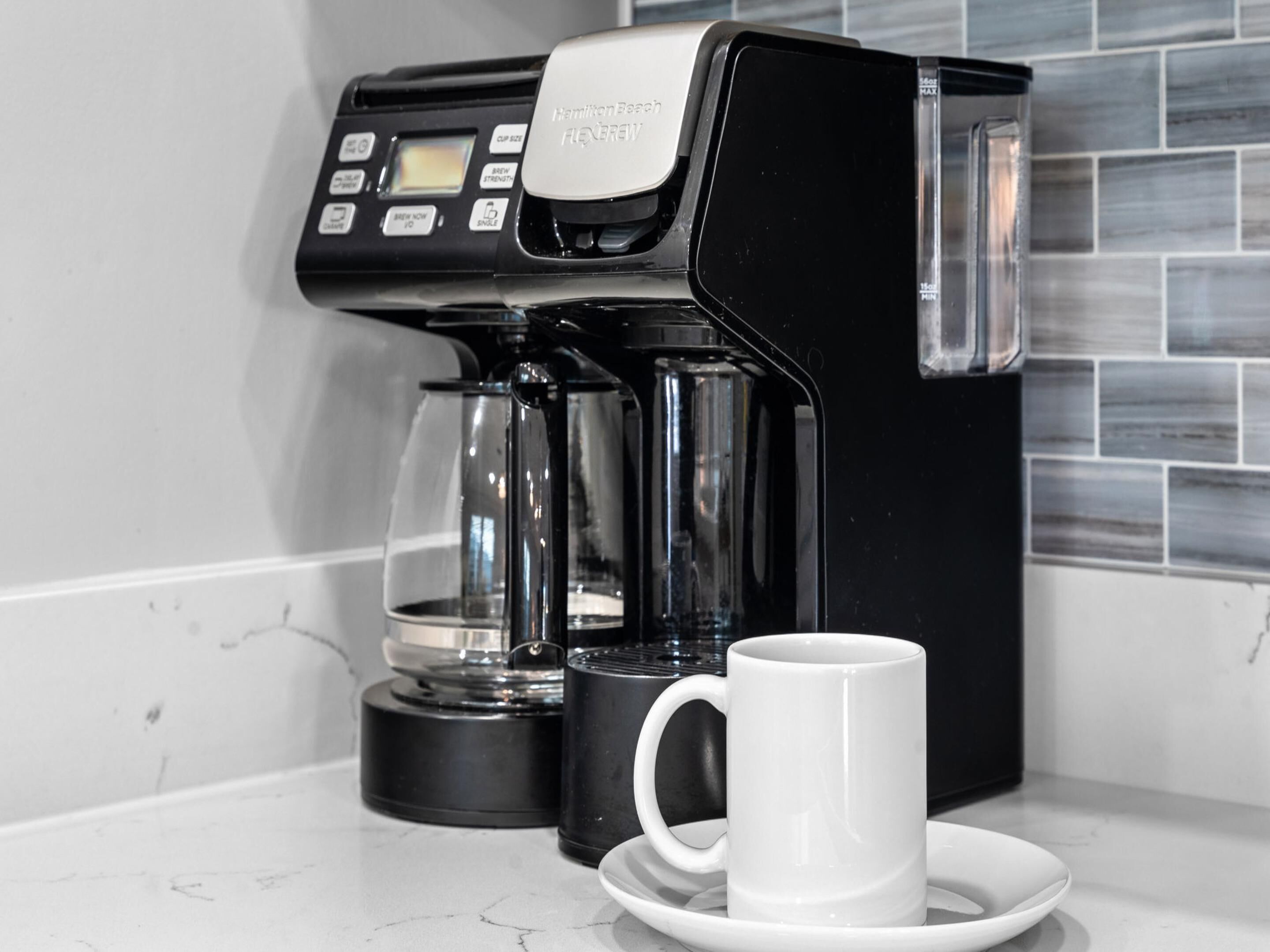 Een zwarte Hamilton Beach Flexbrew koffiezetapparaat met een glazen kan en een witte mok op een wit bord.