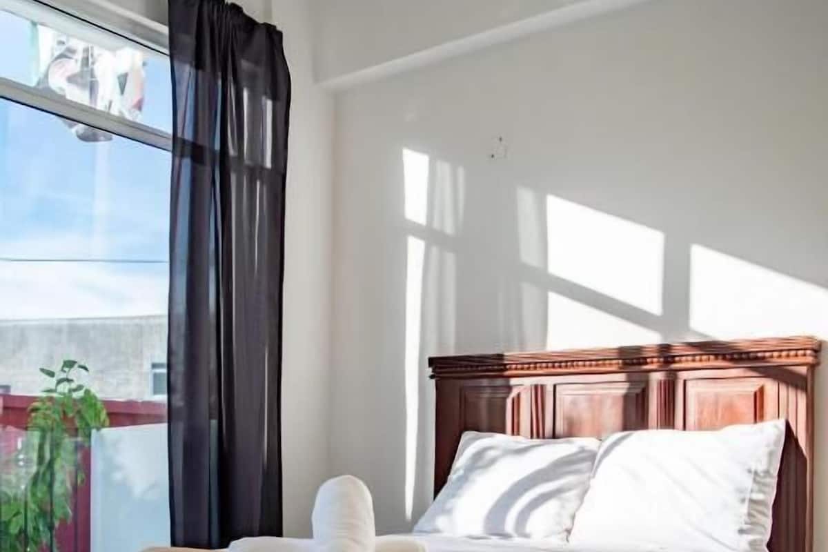 A neatly made bed with white linens and a brown headboard, positioned next to a window with black curtains.