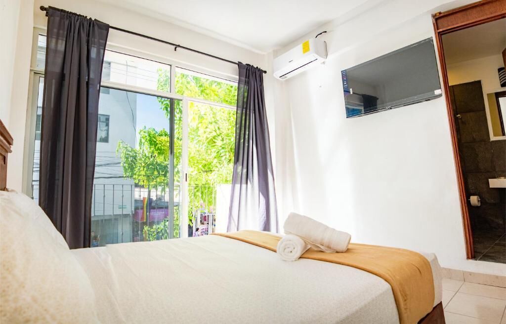 A hotel room with a bed, a window with curtains, a TV, and an air conditioning unit.