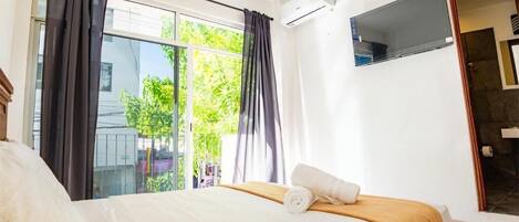A hotel room with a bed, a window with curtains, a TV, and an air conditioning unit.