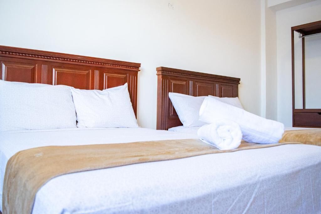 A hotel room with two beds, each with wooden headboards, white bedding, and a rolled towel on the bed.