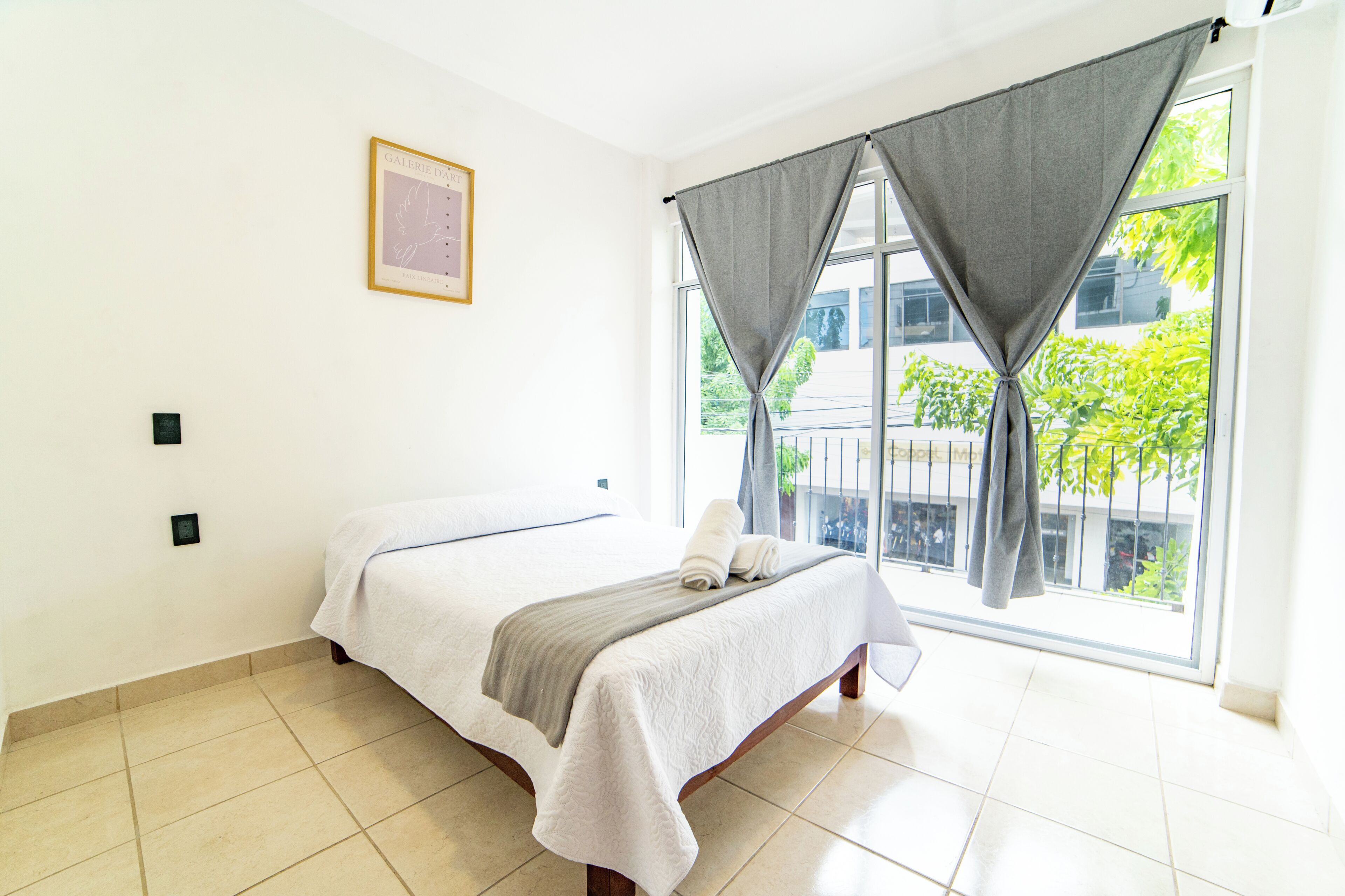 Un dormitorio con cama, ventana con cortinas, un cuadro en la pared y piso de baldosas.