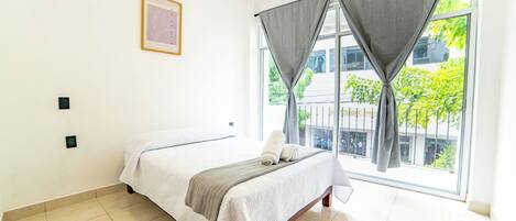 Un dormitorio con cama, ventana con cortinas, un cuadro en la pared y piso de baldosas.
