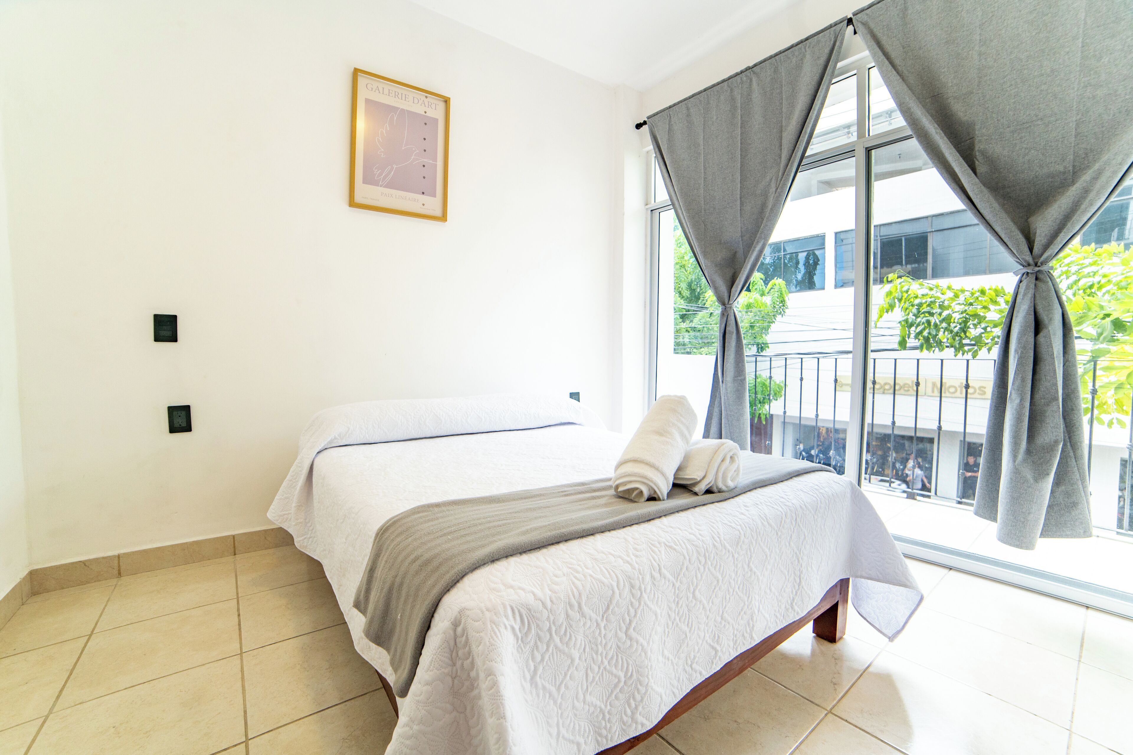 A hotel room with a bed, a window with curtains, a wall-mounted light switch, and a framed picture.