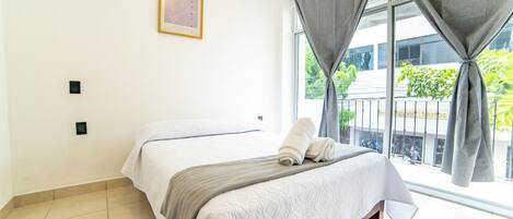 A hotel room with a bed, a window with curtains, a wall-mounted light switch, and a framed picture.