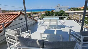 Una terraza en la azotea con mobiliario blanco, incluyendo un sofá y sillones, y una mesa. Hay luces colgantes. La terraza ofrece vistas a una zona costera con edificios y el mar al fondo.