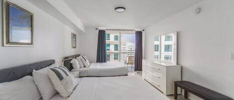 A hotel room with two beds, a dresser, a bench, and a large window with a view of buildings.