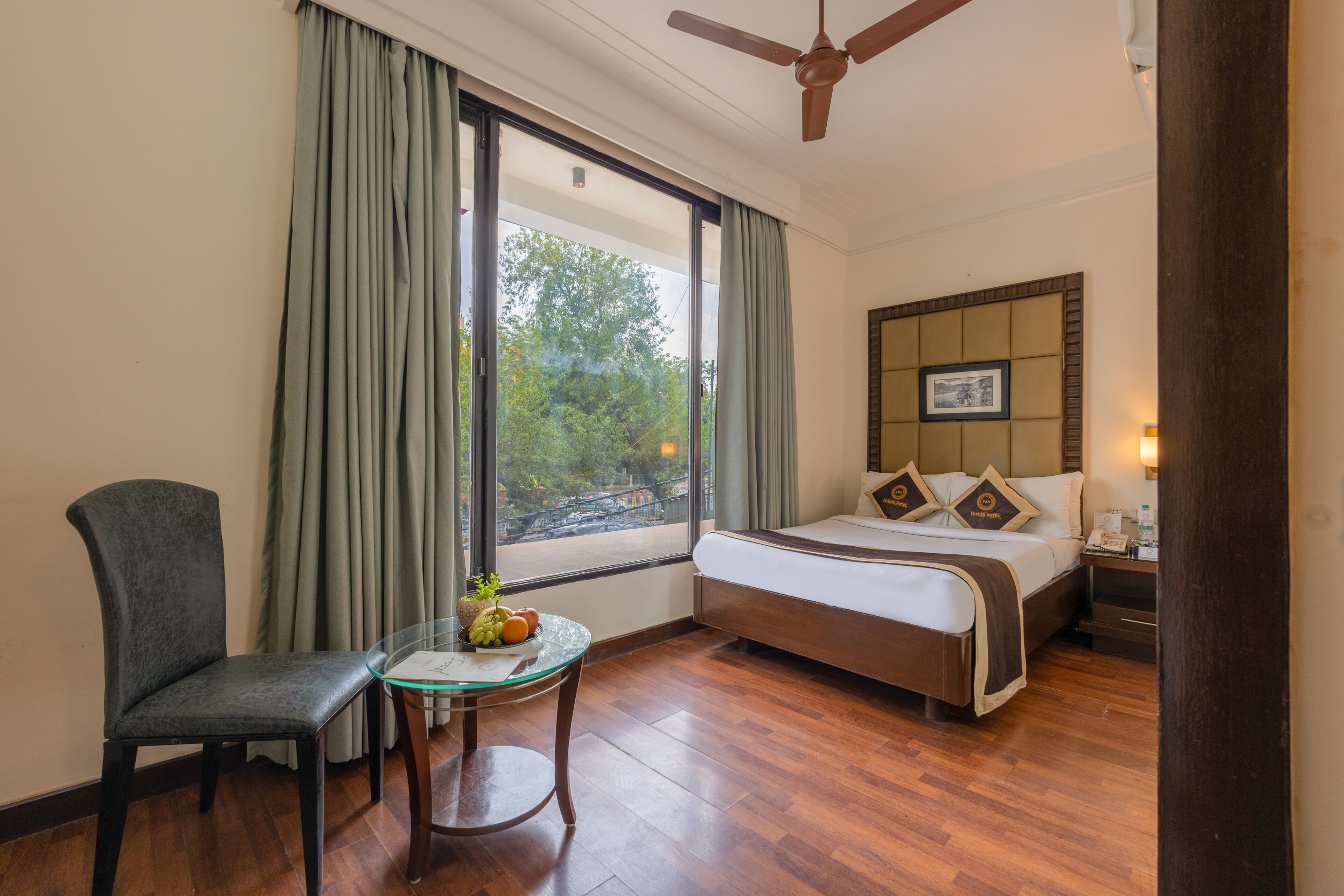 Habitación de hotel con cama, una silla, una mesita con fruta, ventana con cortinas y ventilador de techo.