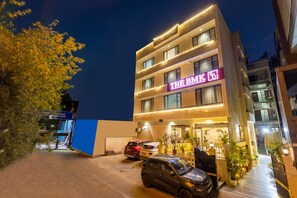 A multi-story hotel with a neon sign, parked cars, and potted plants. - Foxoso The BMK Hotel (New Delhi)