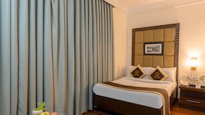 Ein Hotelzimmer mit einem Bett, einem Deckenventilator, einem Nachttisch mit Lampe, einem Glastisch mit Obst und einem Fenster mit Vorhängen.