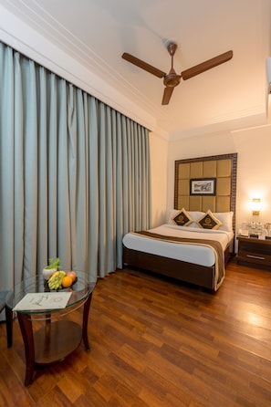 A hotel room with a bed, a ceiling fan, a bedside table with a lamp, a glass table with fruit, and a window with curtains. - Foxoso The BMK Hotel (New Delhi)