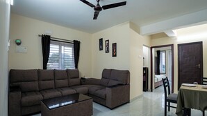 A living room with a sectional sofa, a coffee table, a dining table with chairs, a ceiling fan, and a window with curtains.