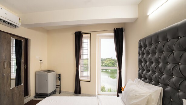 A hotel room with a bed, a window with curtains, a view of greenery, and an air conditioning unit.
