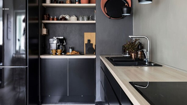 Una cocina moderna con un refrigerador negro, una cafetera y un lavamanos con grifo negro. Hay estantes con diversos utensilios de cocina y una planta en maceta sobre la encimera.