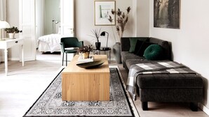Una sala moderna con una mesa de centro de madera, un sofá gris con un cojín verde, un sillón negro y un escritorio blanco con una planta.