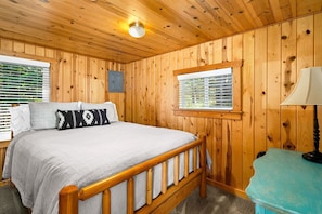 Habitación con paneles de madera, una cama, un ventanal con persianas, una lámpara y una mesita.
