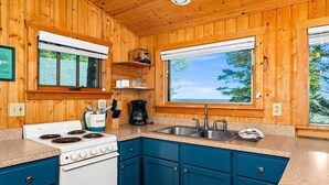A kitchen with wooden walls, a white stove, blue cabinets, and a large window with a view of trees.