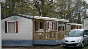 A mobile home with a covered porch, a car parked in front, and a wooded background. - ZEcamping 3 étoiles toboggans dog friendly animations mobil-home TV (Chateauroux)