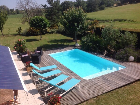 Une piscine rectangulaire dotée d’une terrasse en bois, entourée de verdure et offrant une vue sur un champ herbeux.