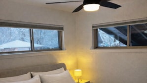 Un dormitorio con cama, una mesita de noche con lámpara, ventilador de techo y ventanas con vistas a árboles.
