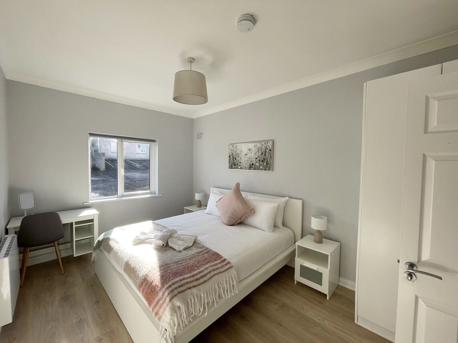 A bedroom with a bed, bedside tables, a desk, a chair, a window, and a door.