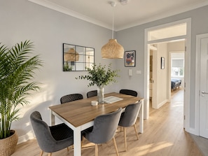 Un coin repas avec une table en bois, des chaises à revêtement gris, un luminaire suspendu en fibres tressées et une plante en pot.