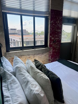 Hotellihuone, jossa on suuri ikkuna, valkoisilla ja vihreillä tyynyillä varustettu sänky sekä punatiilinen seinä.
