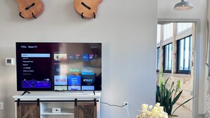 Una sala de estar con un televisor montado en la pared, una mesa de centro de madera con libros y un florero, y dos guitarras colgadas en la pared.