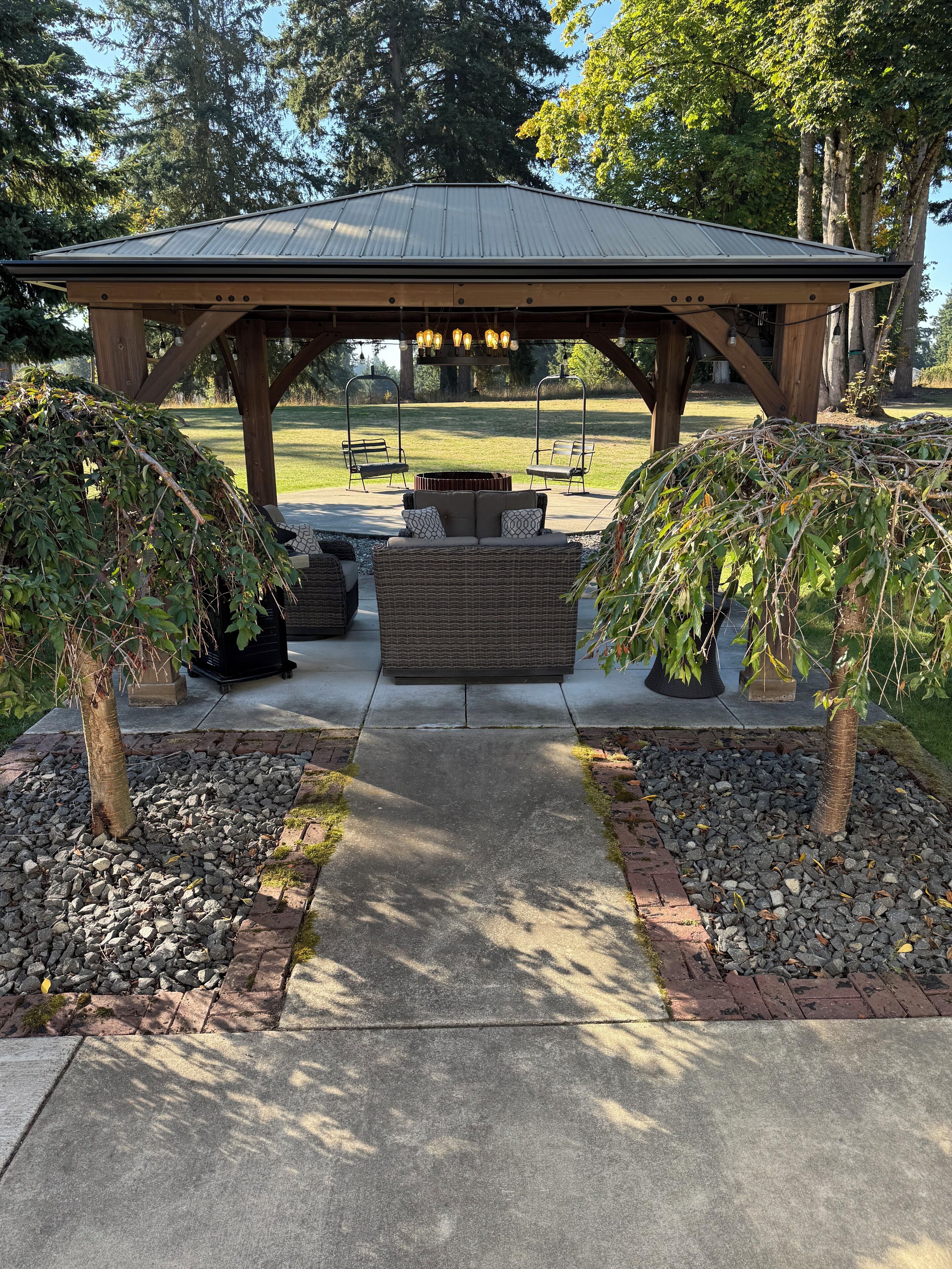 A covered outdoor seating area with a central fire pit, surrounded by stone pathways and small trees.