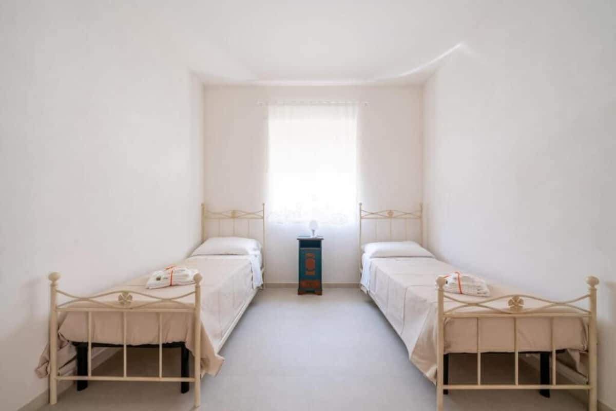 A small, single-room hotel room with two single beds, a small table, and a window with a sheer curtain.