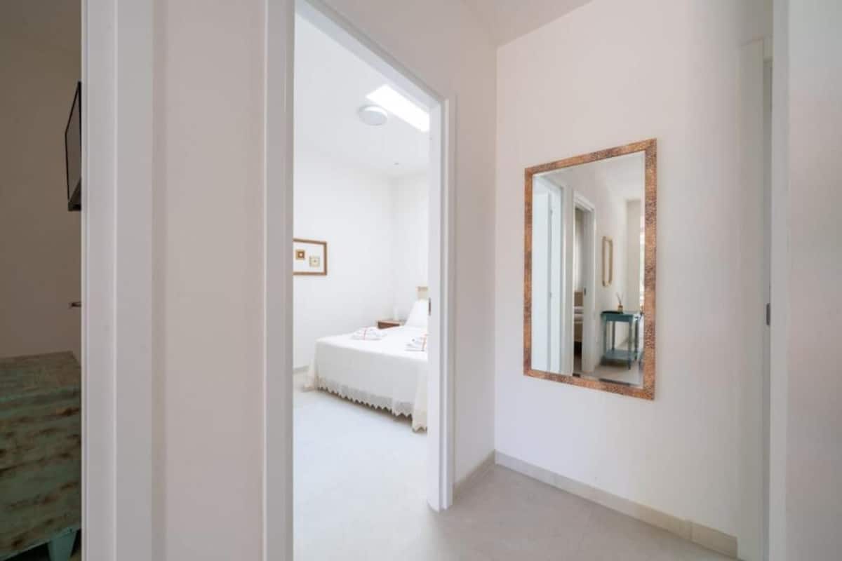 A hallway with a bed in the background, a mirror on the wall, and a door leading to another room.