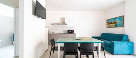 A compact living space with a dining table, four chairs, a blue sofa, a kitchen area with a microwave, and a flat-screen TV mounted on the wall.