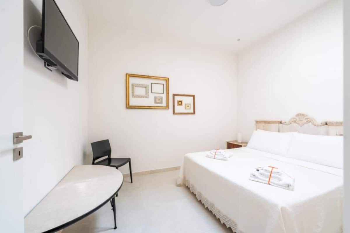 A hotel room with a bed, a desk, a chair, and two framed pictures on the wall.