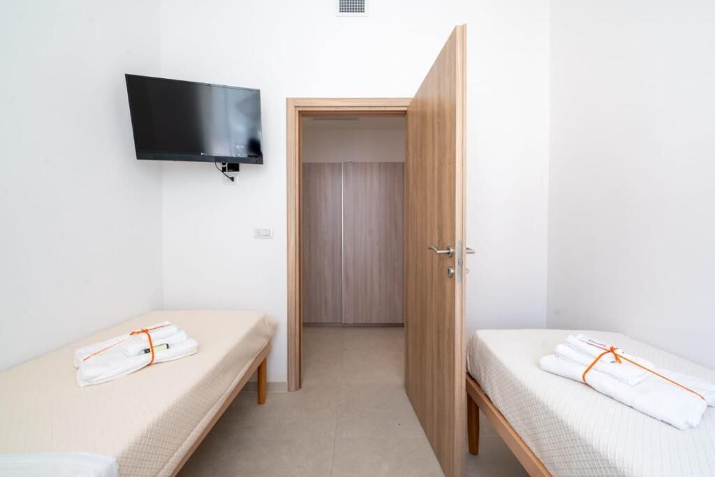 Una habitación pequeña y limpia con dos camas individuales, un televisor fijado a la pared y un armario de madera.