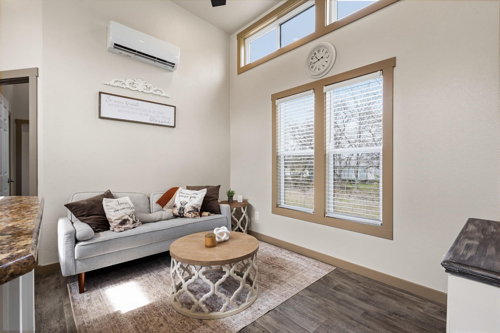 A living room with a grey sofa, a round wooden coffee table, and a large window with blinds.
